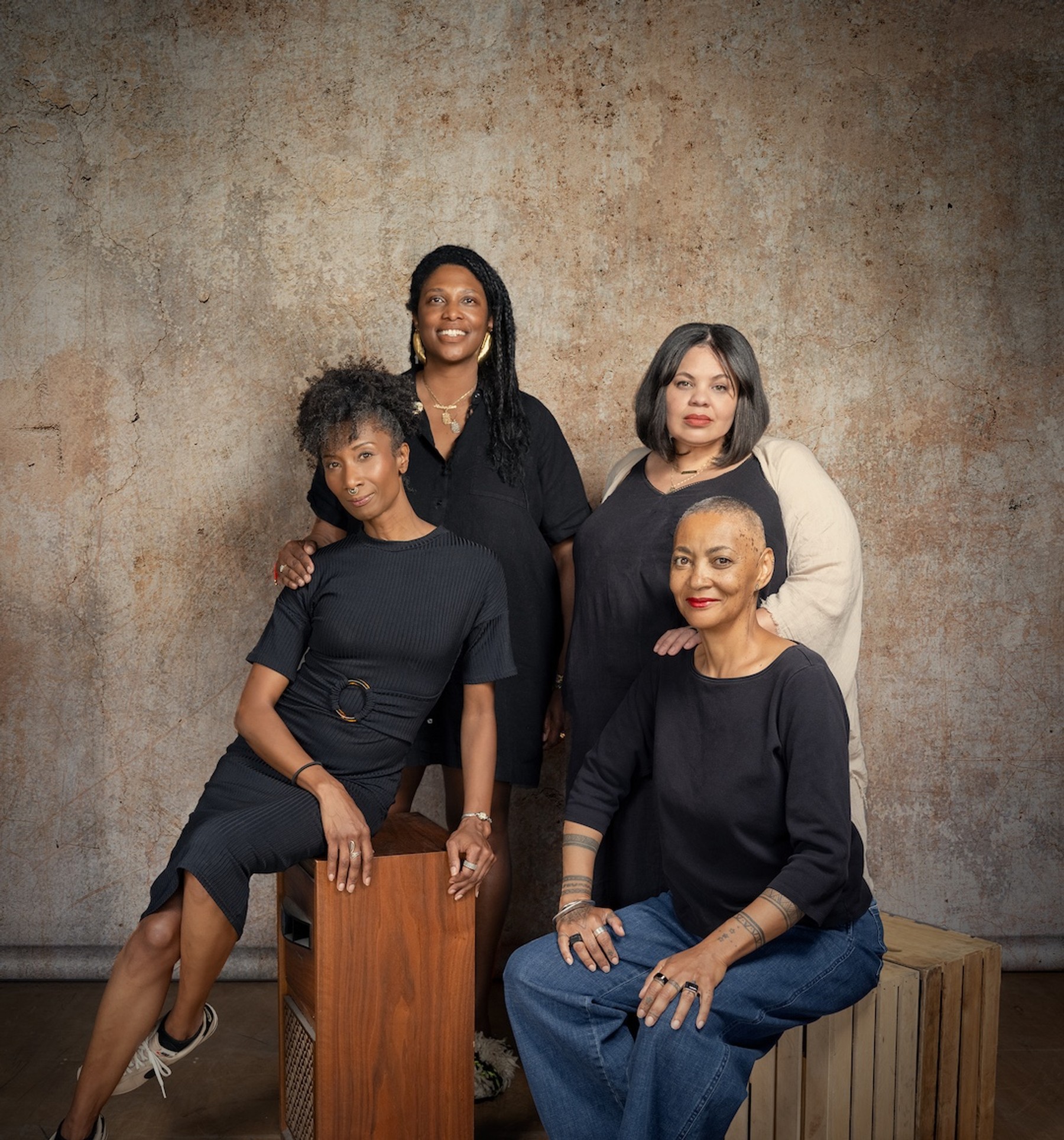 A studio portrait of four African American women dressed in black, posed together against a mottled brown backdrop. Two are seated on wooden crates in front, and two stand behind them, resting their hands on the others' shoulders. The composition is close and relaxed, conveying both individuality and collective presence.