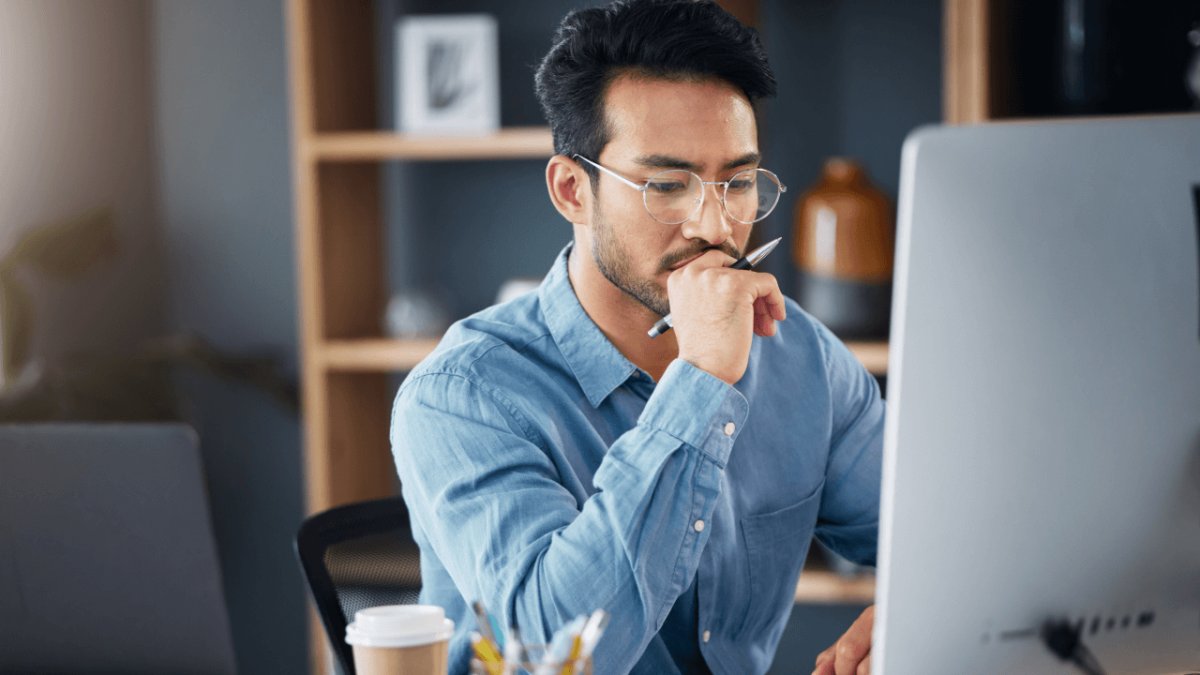 A man in glasses looks thoughtfully at a computer screen, holding a pen to his chin.