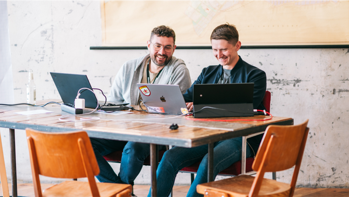 Two men smiling while working on laptops at a table.