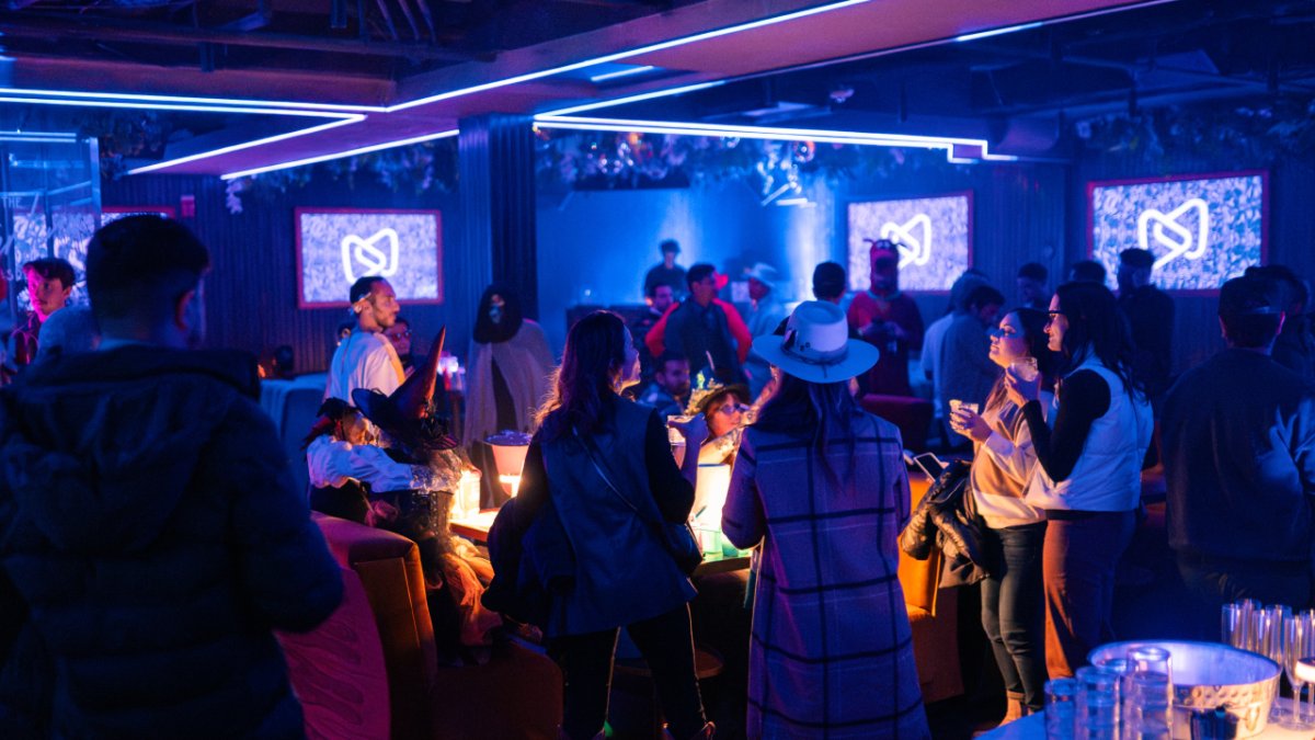 People socialize in a dimly lit club under blue neon lights, with screens showing a white logo.