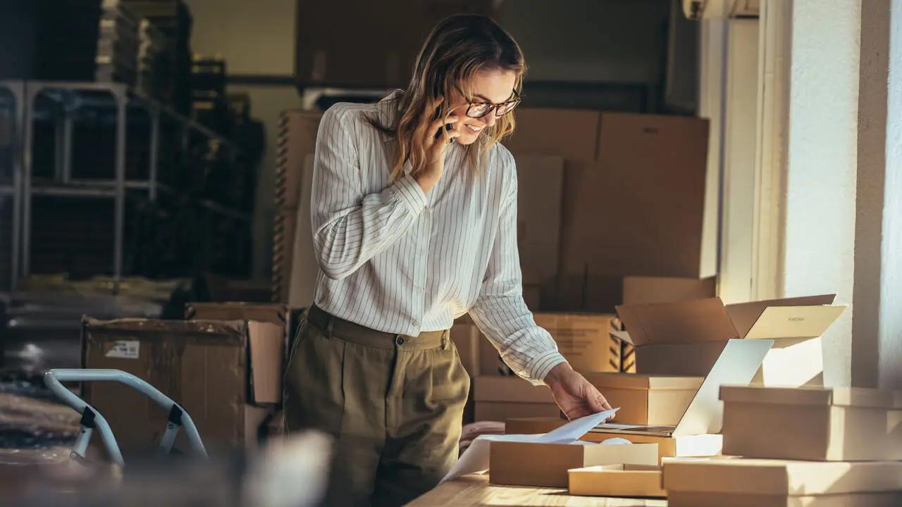 Smiling woman on phone sorting packages in a fulfillment center.