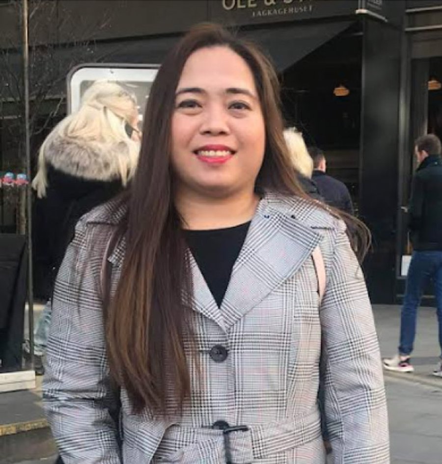 Una donna sorridente con capelli lunghi castani indossa un cappotto grigio a quadri all'aperto.