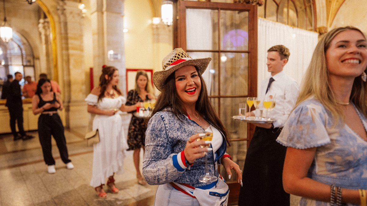 A smiling woman in a straw hat and denim jacket holds a drink at an indoor event, with other guests and a server in the background.