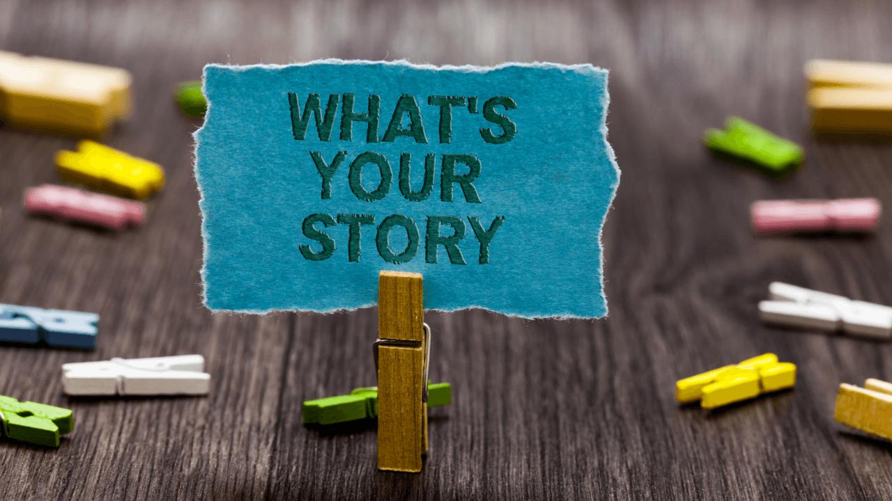 Blue paper with "WHAT'S YOUR STORY" held by a clothespin on a wooden background with colorful mini clothespins.