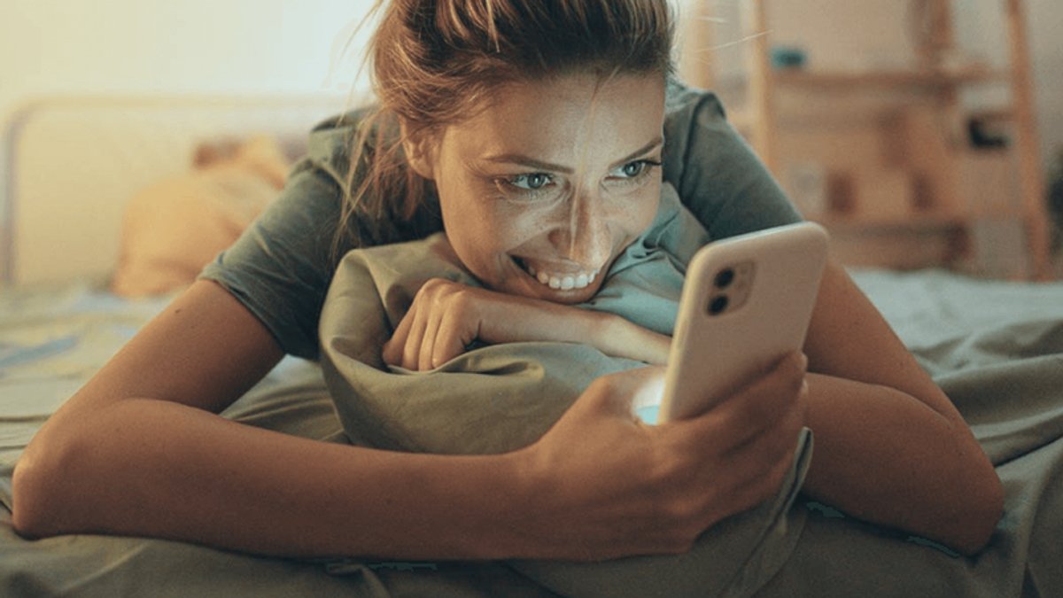 A smiling woman lies on her stomach in bed, looking at her glowing phone screen.