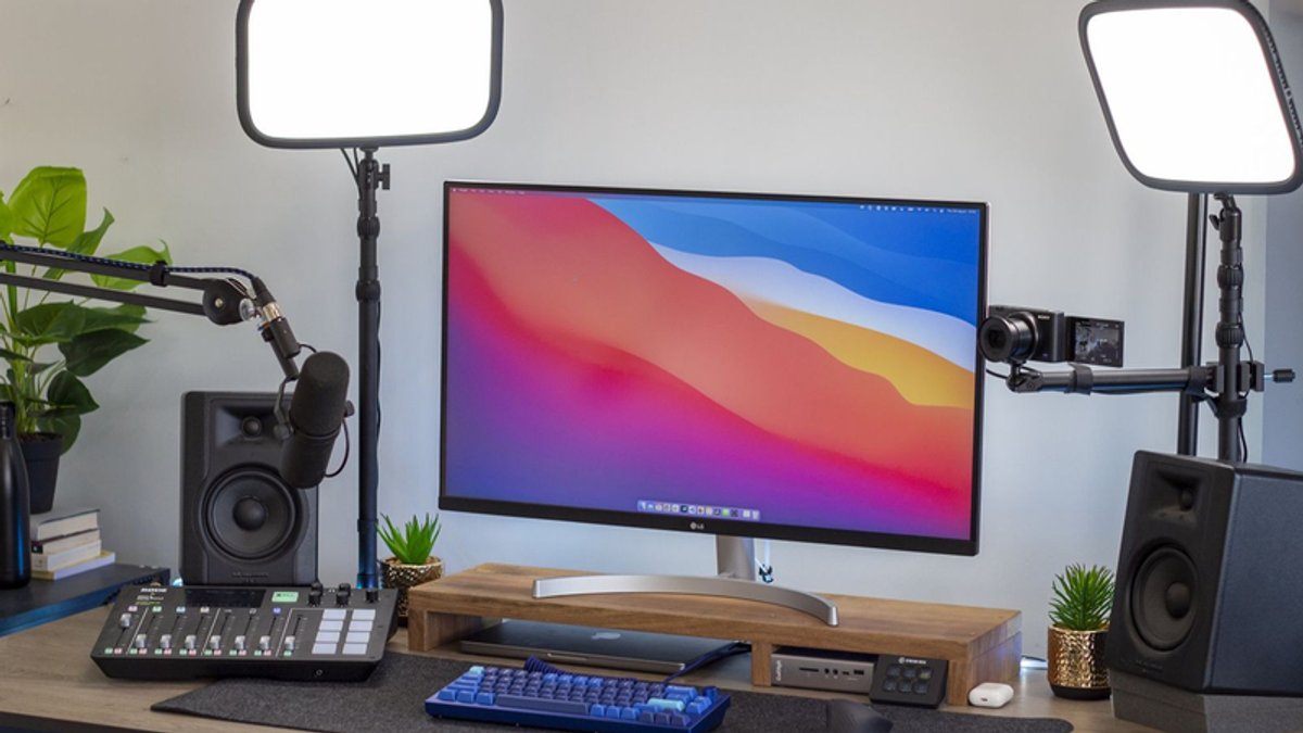 A desk setup featuring a monitor with macOS Big Sur wallpaper, a microphone, camera, two LED panel lights, an audio mixer, and two studio monitors.