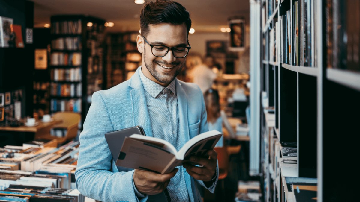 A smiling man wearing glasses reads a book in a bookstore.