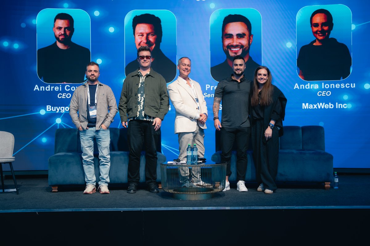 Five panelists, four men and one woman, stand on a stage in front of a blue screen displaying their headshots and professional titles.