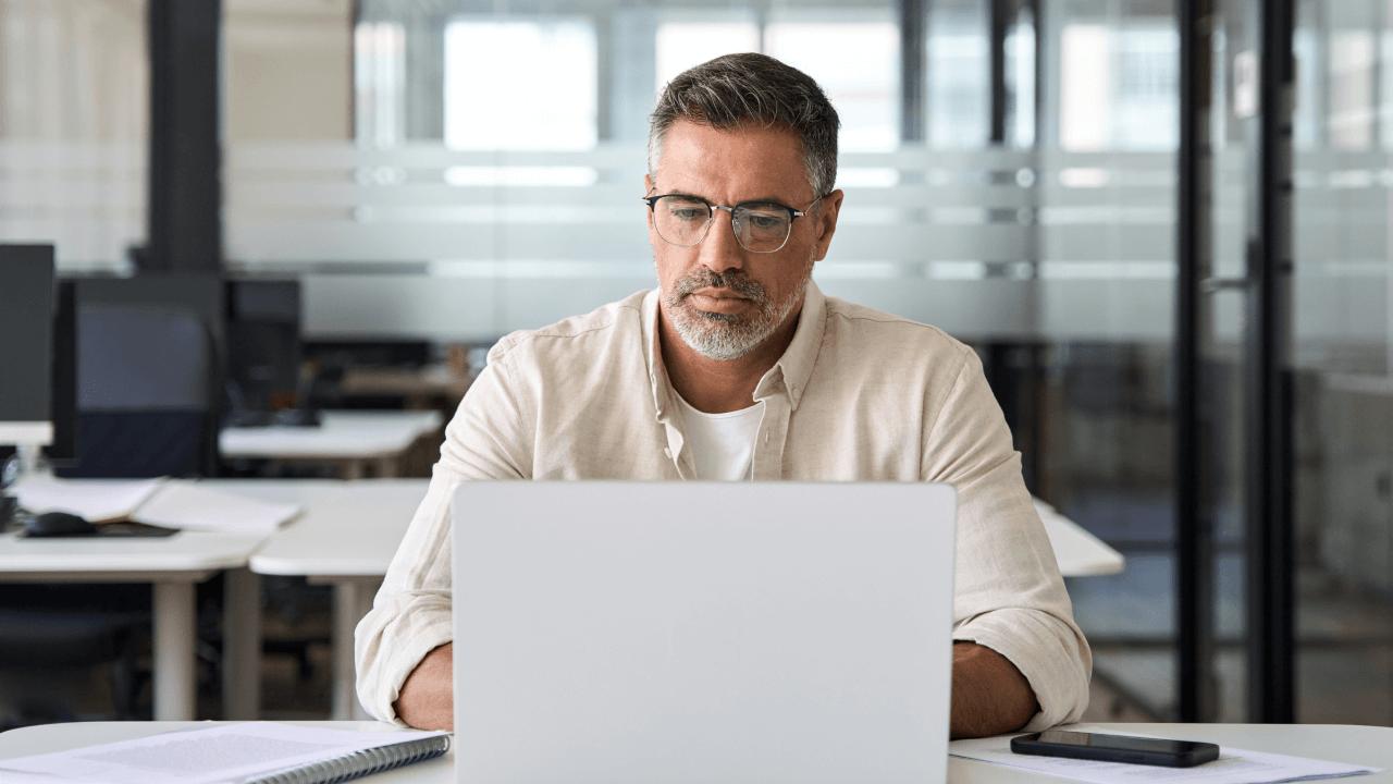 Man with glasses and a beard looking at a laptop in an office.