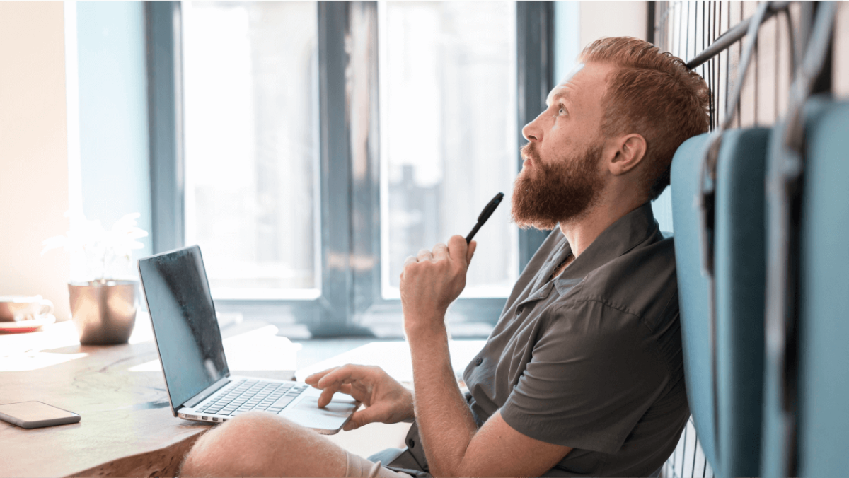 A thoughtful man with a beard holds a pen to his chin while looking up from his laptop.