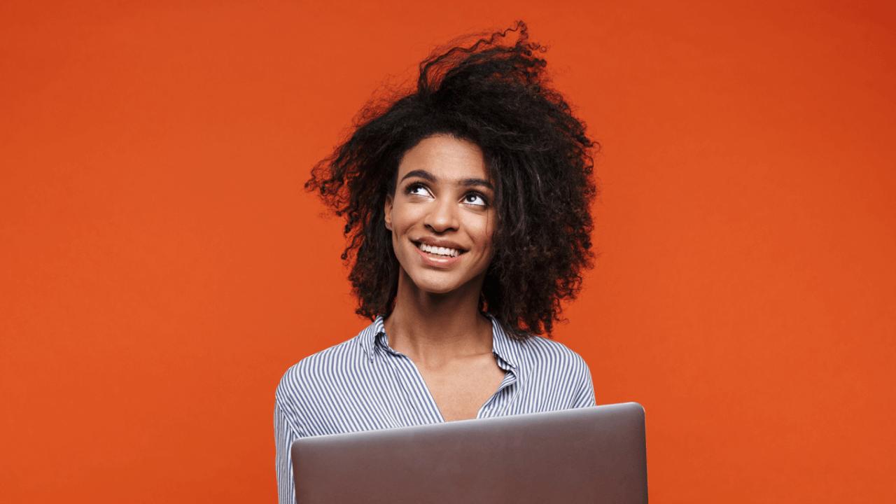 Smiling Black woman with curly hair looks up, holding a laptop against an orange background.