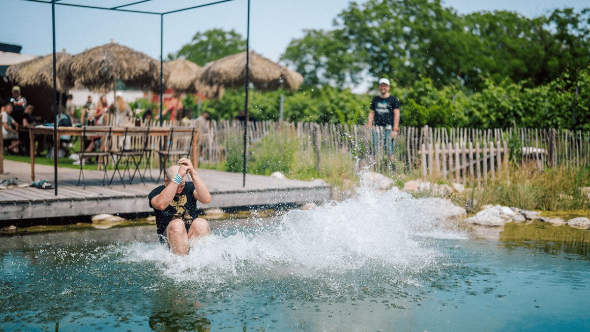 A man cannonballs into a natural pond, creating a huge splash, with onlookers in the background.