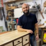 A smiling, bald, bearded man leans on a two-toned wooden cabinet with light, textured drawers and a dark frame, in a blurred workshop.