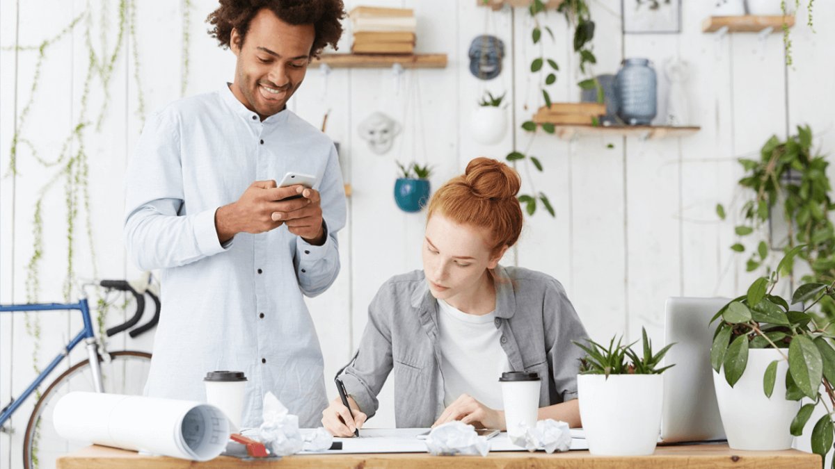 A man stands smiling at his phone next to a woman writing at a desk, surrounded by plants.