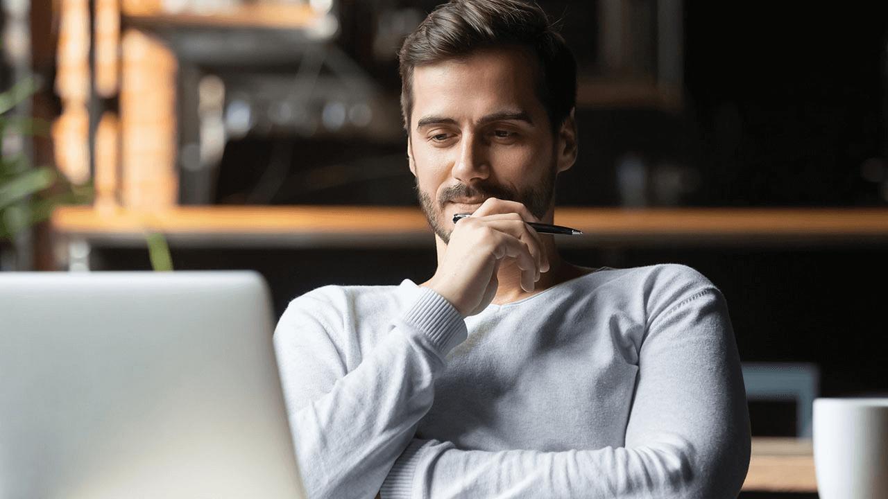 Man in a light sweater looking at a laptop, holding a pen to his chin in thought.