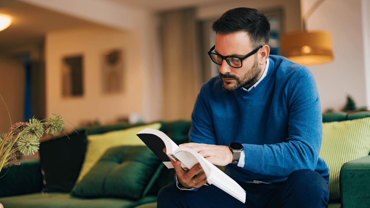 A man in a blue sweater and glasses sits on a green couch, reading a book.
