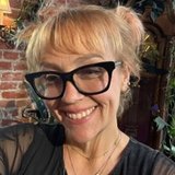 A smiling woman with blonde pigtails and black glasses wears a black top and layered necklaces against a brick wall with plants.