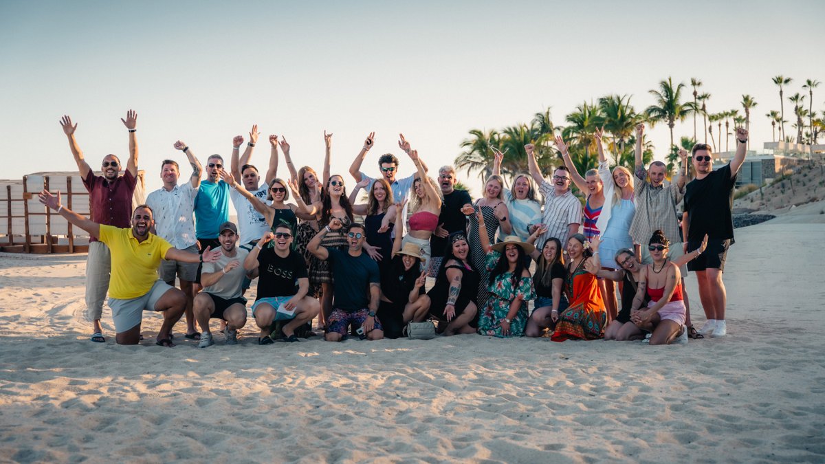 A large group of excited people on a beach with arms raised and palm trees in the background.