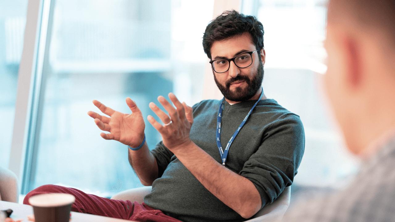 A bearded man wearing glasses and a blue lanyard gestures with open hands while talking to another person.