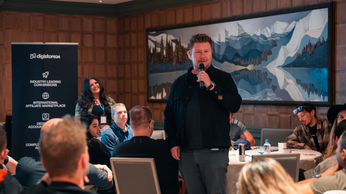 A man speaks into a microphone at a Digistore24 event, addressing an audience seated at tables.