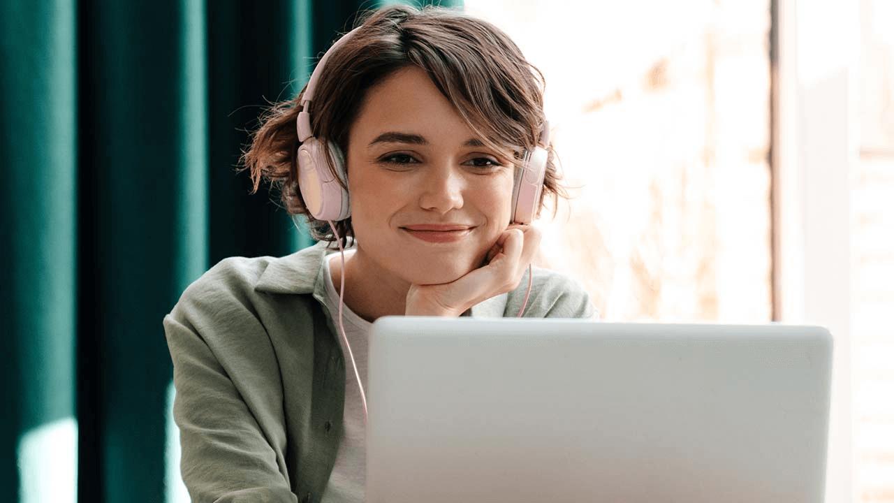 Smiling woman wearing pink headphones, looking at the camera with a laptop in front.