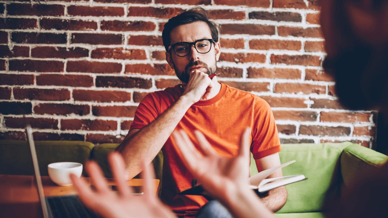 Man with glasses listens intently as another person gestures in the foreground.