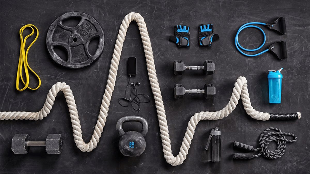 Gym equipment arranged on a dark background, including a wavy battle rope, dumbbells, kettlebell, weight plate, resistance bands, jump rope, gloves, and water bottles.