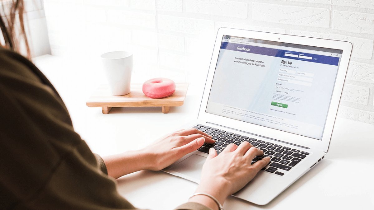 Person typing on a laptop displaying the Facebook signup page, with a pink donut and a white cup on a tray.