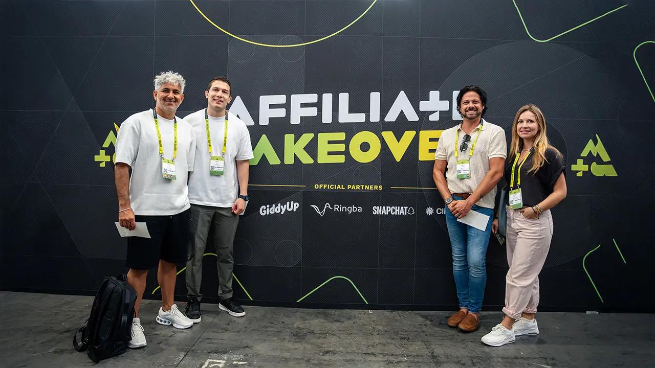 Four people standing and smiling in front of a black wall with "AFFILIA+ TAKEOVER" text and partner logos.