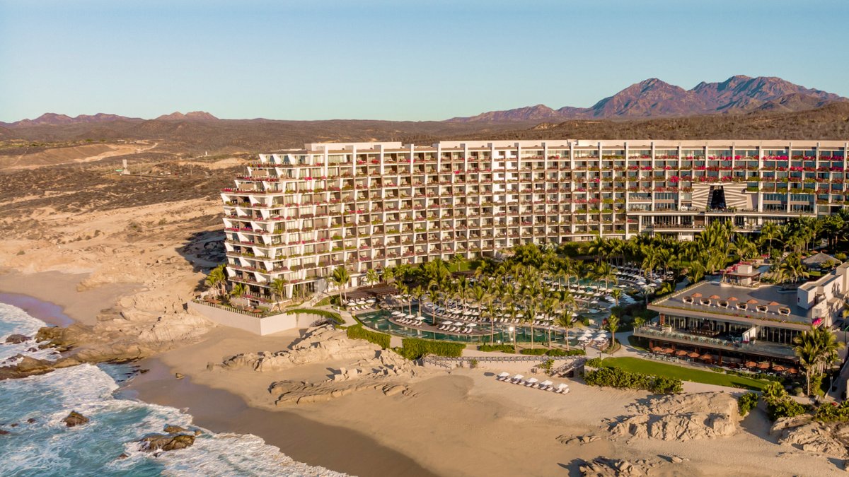 Aerial view of a large, modern beachfront resort with pools, palm trees, and distant mountains.