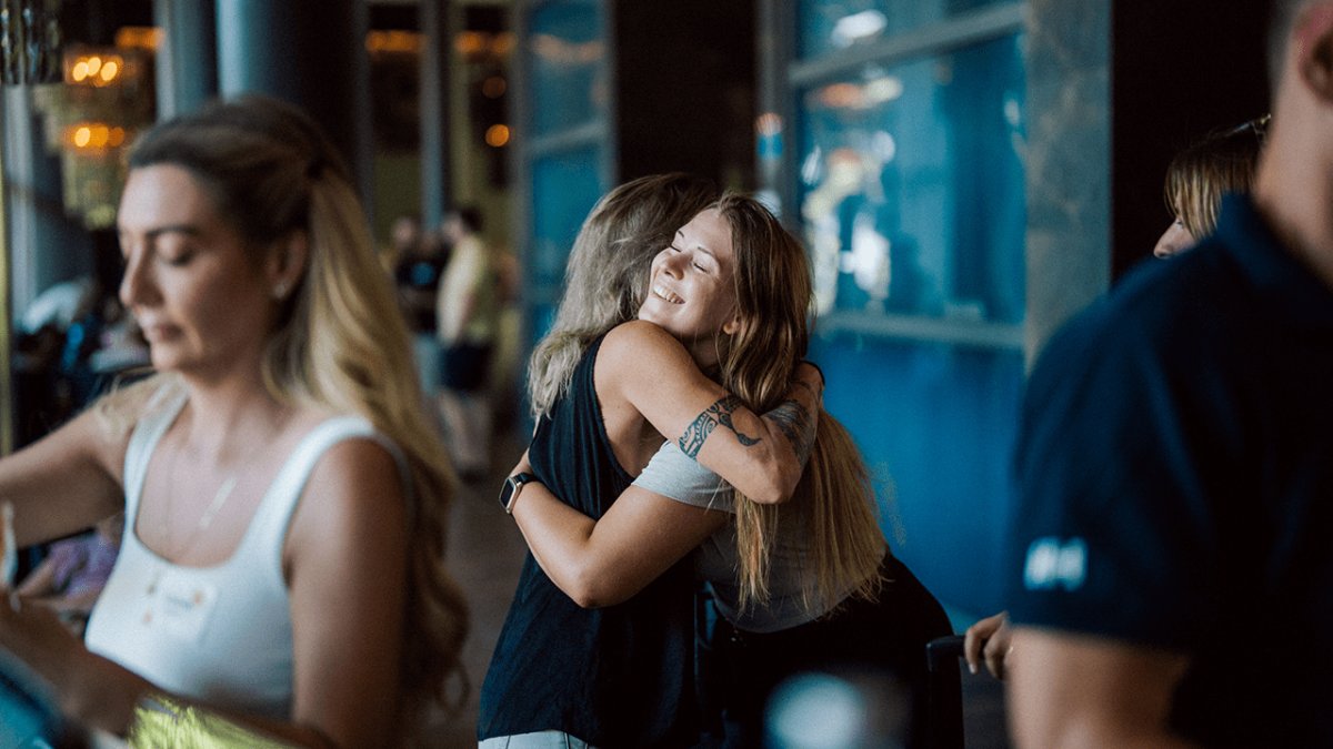 Two smiling women hug each other warmly in a busy room.