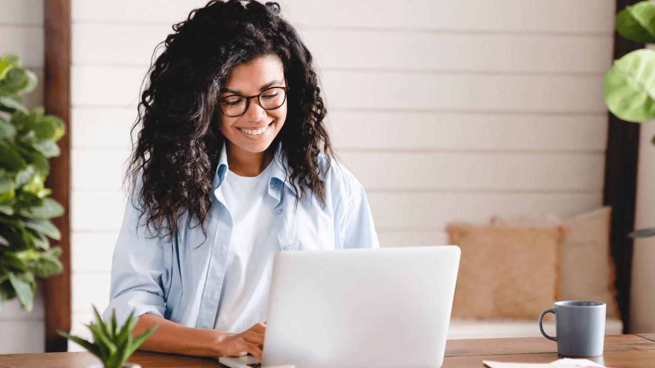 A smiling woman with dark curly hair and glasses works on a laptop at a desk.