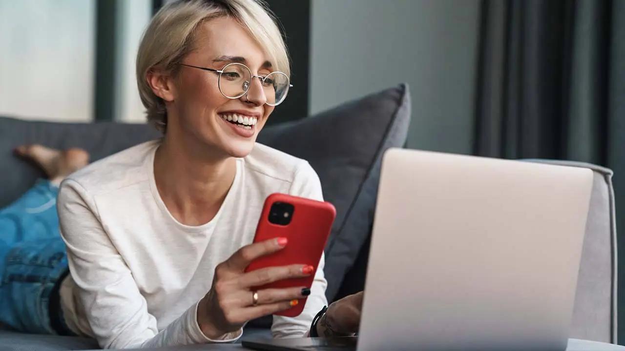 A smiling woman with glasses lies on a couch, holding a smartphone and using a laptop.