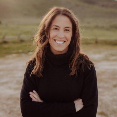 A smiling woman with long brown hair wears a black turtleneck sweater, arms crossed, standing outdoors.
