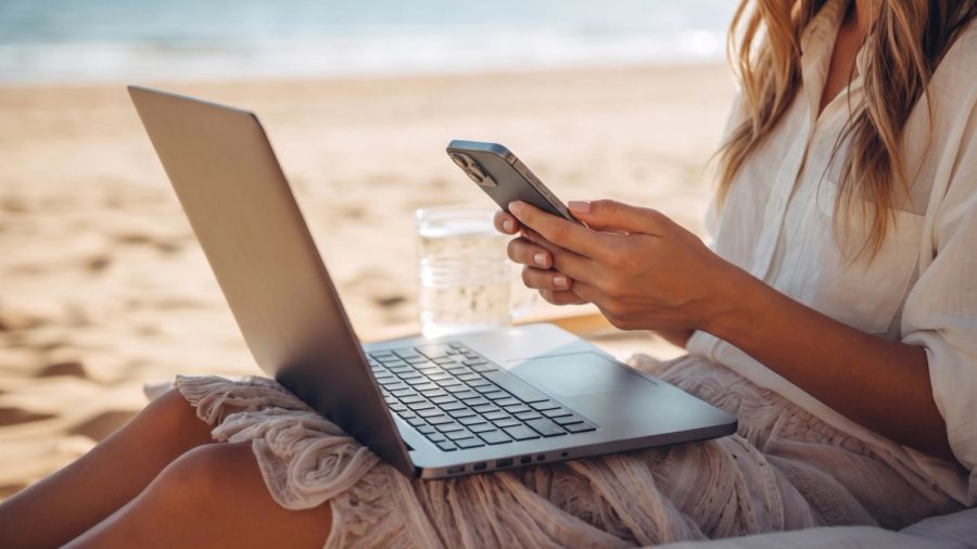 Woman working on a laptop and smartphone on a beach.