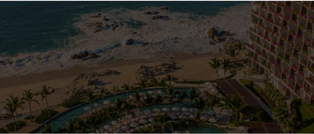 Aerial view of a resort with a hotel, swimming pool, palm trees, sandy beach, and ocean waves.