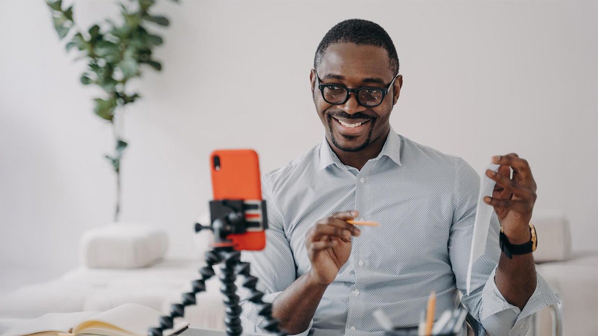 A smiling Black man with glasses records a video on a smartphone, holding a pen and paper.