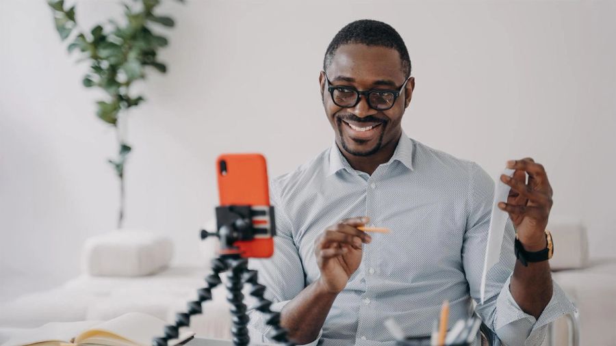 A smiling Black man with glasses records a video on a smartphone, holding a pen and paper.