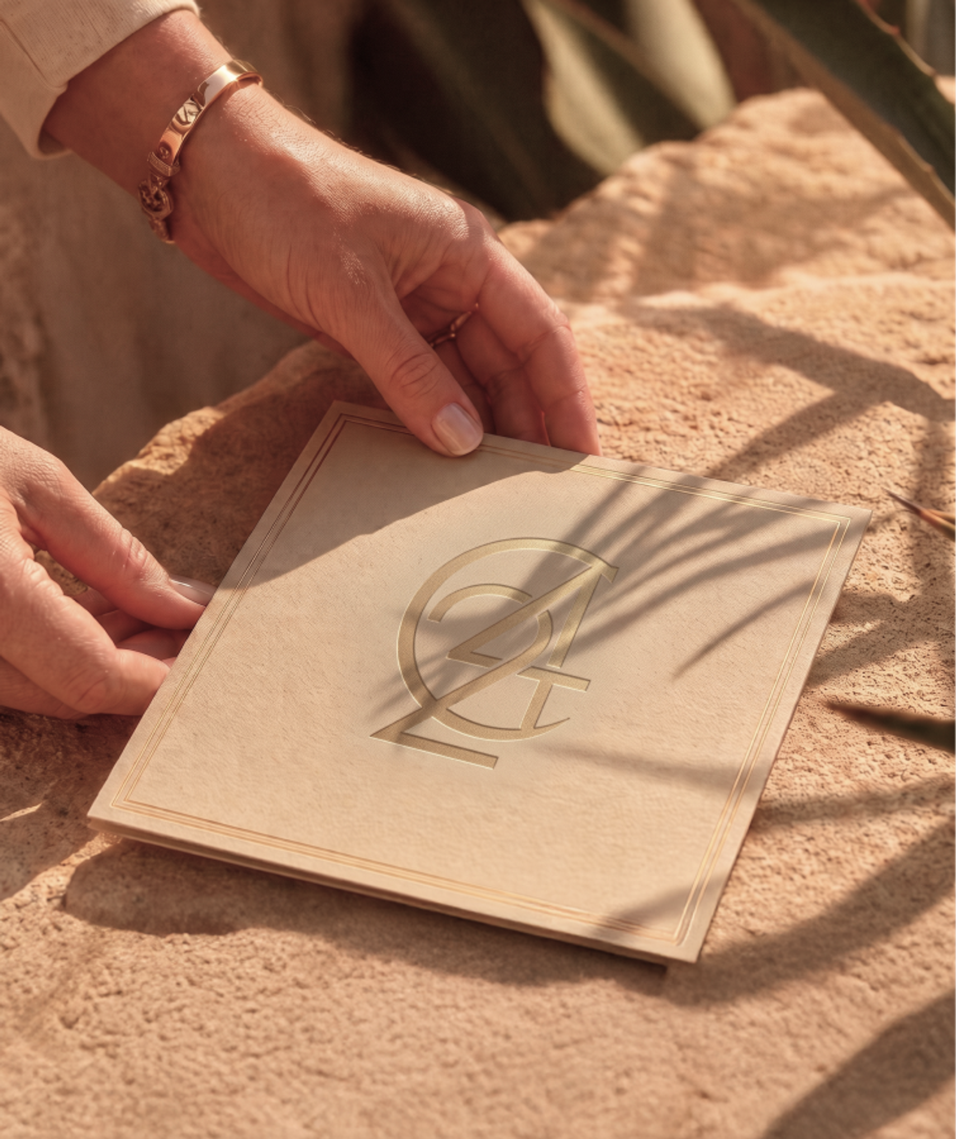 Hands gently hold a square tan card with an embossed logo, resting on a textured surface with leaf shadows.