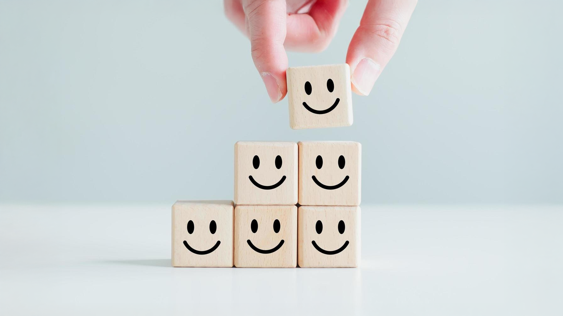 A hand places a smiley face wooden block on top of a pyramid of other smiley face blocks.