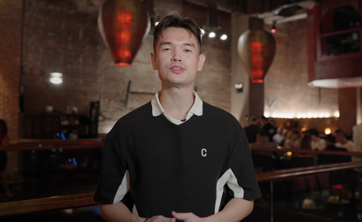 A young man in a black and white polo shirt with a 'C' logo stands in a dimly lit restaurant with large red light fixtures.