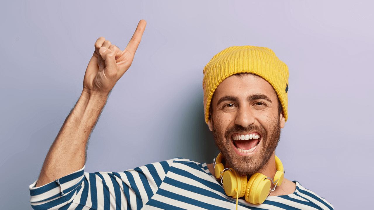 A happy man in a yellow beanie and headphones points upwards.