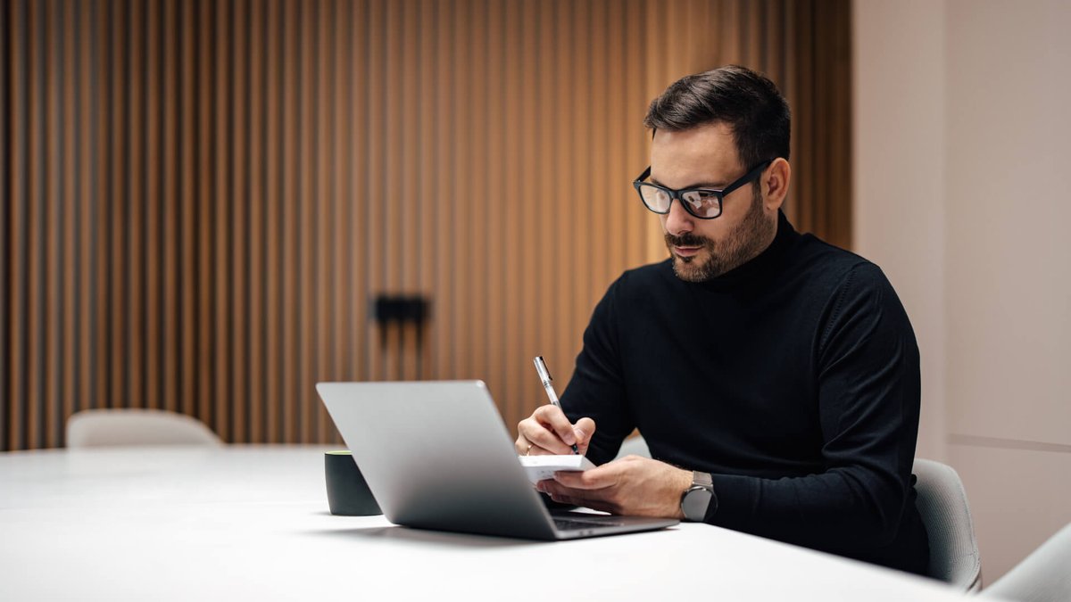 A man in glasses and a black turtleneck writes in a notebook with a laptop nearby.