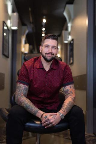 A smiling, bearded man with tattooed arms sits on a stool, wearing a red patterned shirt.