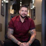 A smiling, bearded man with tattooed arms sits on a stool, wearing a red patterned shirt.