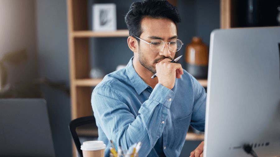 A man in glasses looks thoughtfully at a computer screen, holding a pen to his chin.