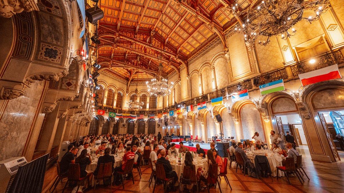 An ornate banquet hall filled with people dining, decorated with international flags and chandeliers.