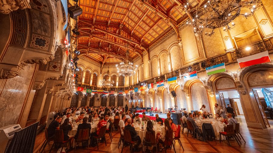 An ornate banquet hall filled with people dining, decorated with international flags and chandeliers.