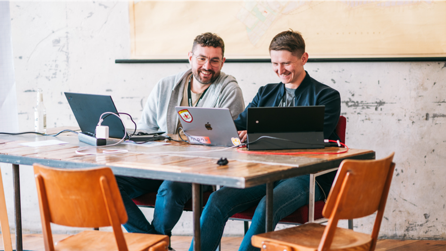 Two men smiling while working on laptops at a table.