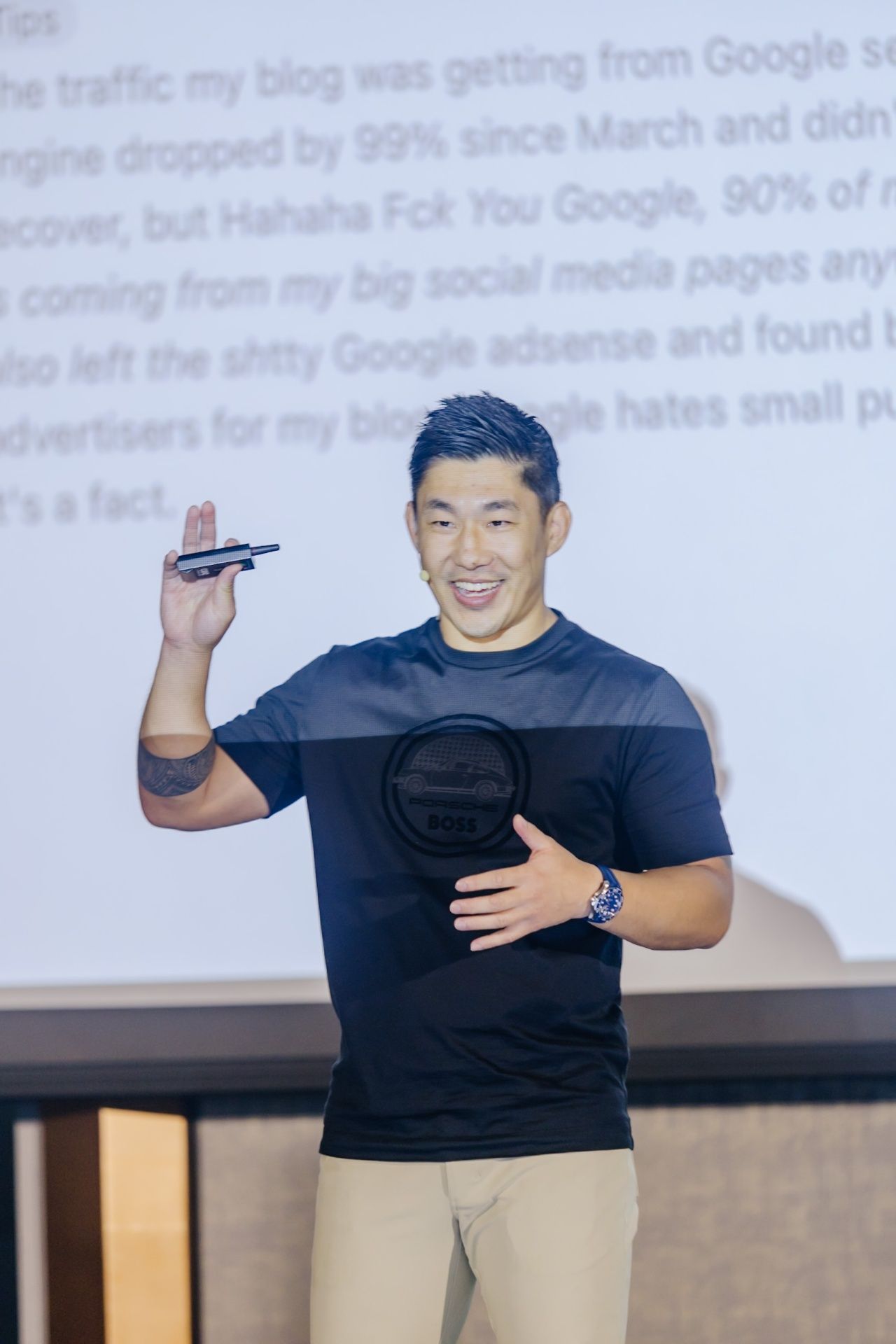 A smiling Asian man in a dark t-shirt holds a clicker while presenting in front of a screen with blurred text.